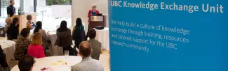 Attendees gather attentively around a speaker at the UBC Knowledge Exchange event, with a banner banner stated "UBC knowledge Exchange. We build a culture of knowledge of knowledge exchange through training, resources, and tailored supports for the UBC research community" 