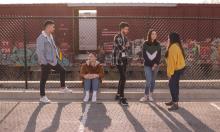 Photo of 5 young adults standing in an industrial area, in front of a chain link fence, copyright by Eliott Reyna on Unsplash